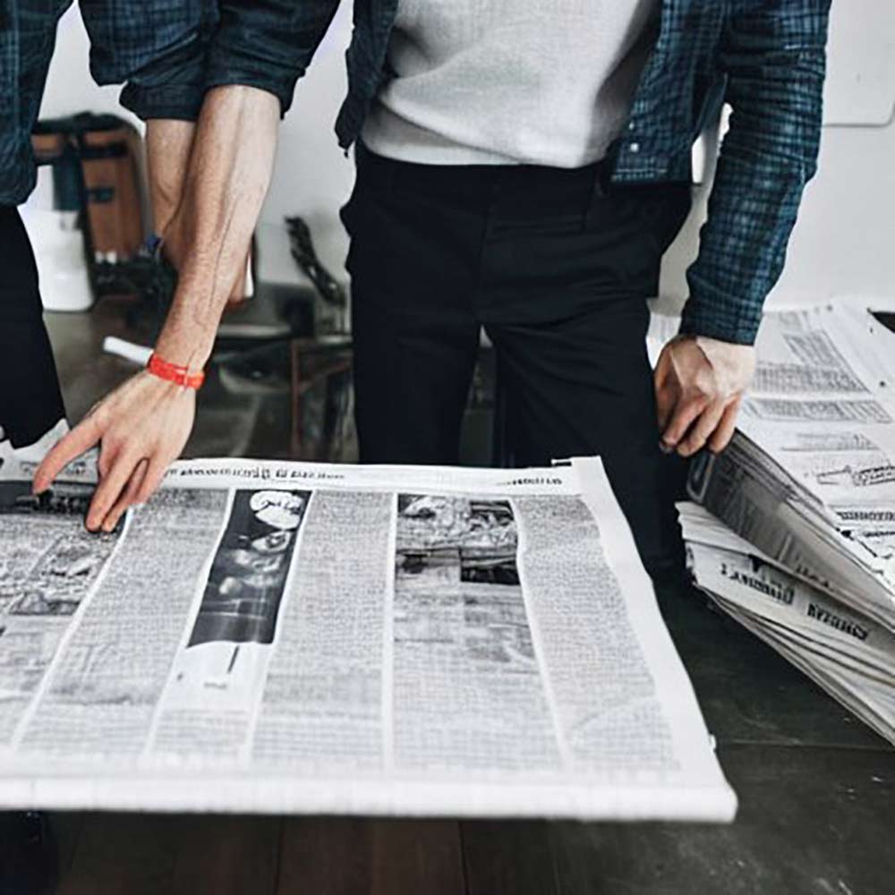 KI generiertes Bild zweier Personen, von denen aber nur der Torso im Bild ist, die gemeinsam eine Zeitung auf einem Tisch ausbreiten. Das Bild zeigt die typischen Fehler von KI generierten Bildern: Die Hand der von uns aus rechten Person hat sechs ziemlich deformierte Finger, der Arm der von uns links stehenden Person ist in den Arm der rechtsstehenden Person deformiert.