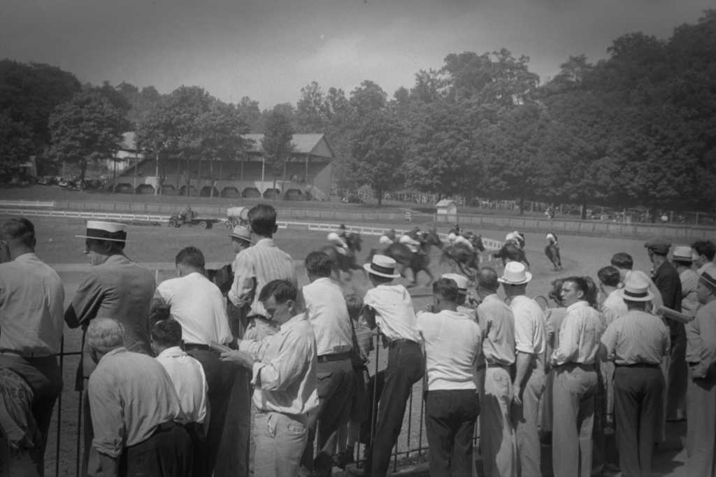 Foto aus den USA, aus dem Jahr 1938, schwarz-weiß: Eine größere Menge Zuschauer betrachtet ein Pferderennen in Lancaster, Ohio. Wir sehen ausschließlich Männer, etwas schräg von hinten fotografiert, aufgereit an einer Balustrade, gekleidert in hellen Sommerhosen und meist weißen Hemden, deren Kragen aufgeknöpft und die Ärmel hochgekrempelt sind, einige tragen Sommerhüte, sogenannte Kreissägen. Im Hintergrund sehen wir das Feld des Pferderennens, mit Jockeys besetzte Pferde auf einer Rennbahn, die gerade an den Zuschauern vorbeigerast sind und sich nun in Richtung Kurve entfernen.
