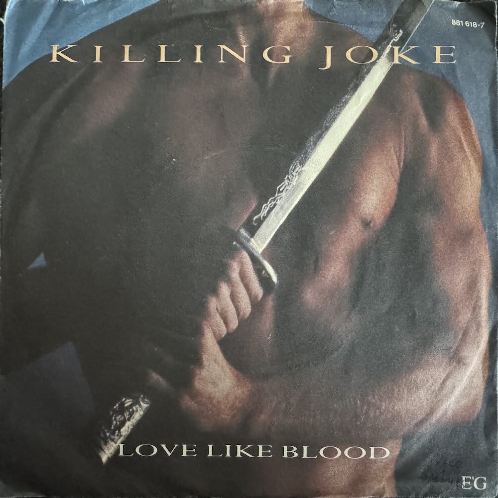 Plattencover, 7inch Single, Killing Joke, Love like Blood. Es ist der Torso eines sehr muskulösen, nackten Mannes zu sehen, der vor seiner Brust diagonal von unten links nach oben recht ein silberglänzendes Samurai-Schwert hält.