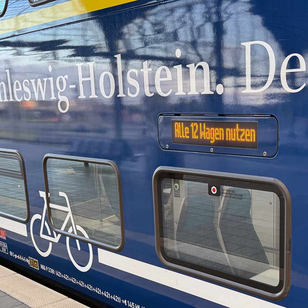 Ein Personenwagen der Regionalbahn Nord im Ausschnitt, der Wagen ist blau mit gelben Dekors, ein Teil der Aufrschrift Schleswig-Holstein, der echte Norden ist zu sehen. Die Anzeige am Wagen zeigt an: bitte alle 12 Wagen nutzen.