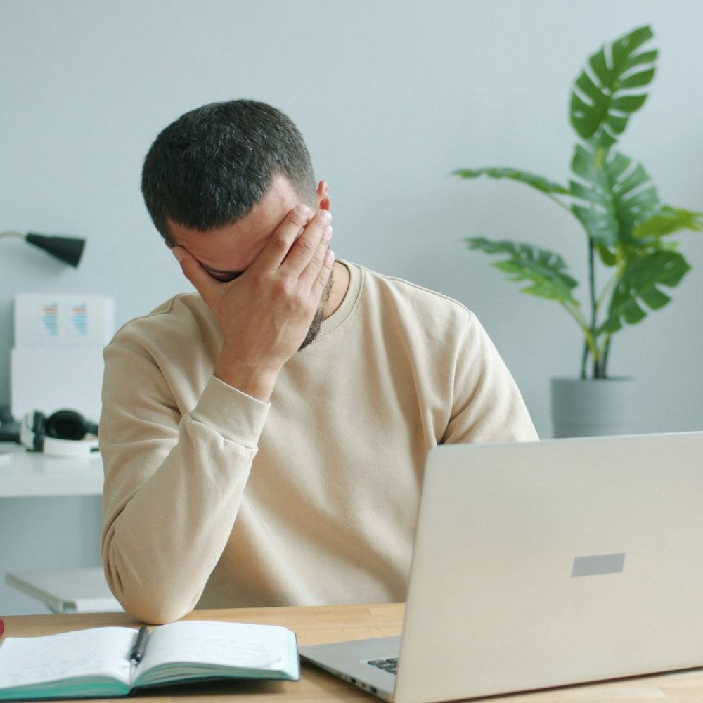Eine Person sitzt an einem Schreibtisch vor einem Laptop und hält sich mit einer Hand das Gesicht, als wirke sie gestresst oder erschöpft. Auf dem Tisch liegen ein Notizbuch, Stifte und ausgedruckte Diagramme; im Hintergrund sind ein Regal, eine Schreibtischlampe und eine Zimmerpflanze zu sehen.