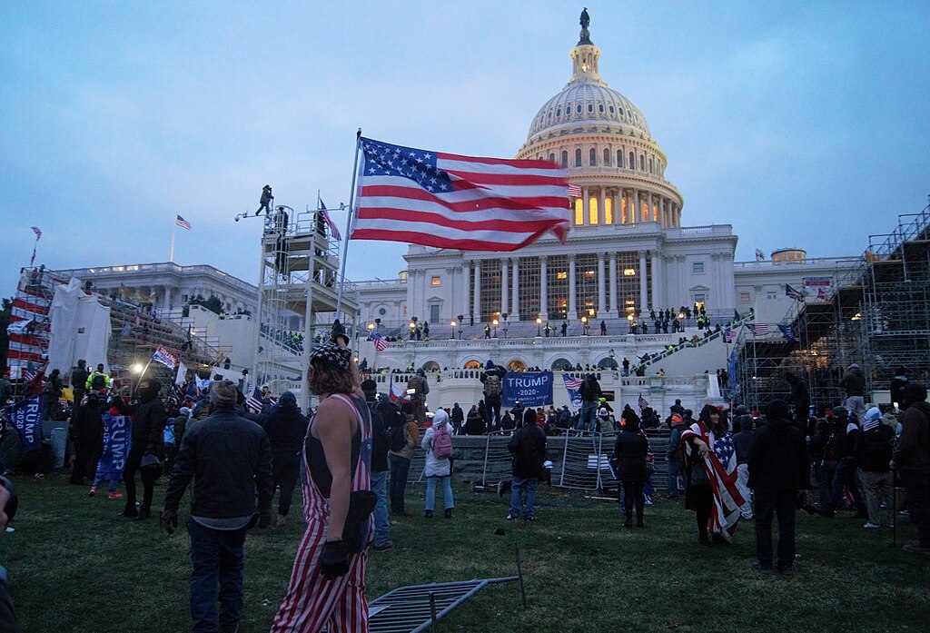 Das Bild zeigt eine große Menschenmenge vor dem weißen, kuppelgekrönten Gebäude des US-Kapitols in Washington, D.C. Es ist Abend oder früher Morgen, das Licht ist kühl und der Himmel leicht bewölkt. Viele Menschen stehen auf dem Rasen und auf den Treppen vor dem Gebäude. Einige tragen amerikanische Flaggen oder sind in Flaggen gehüllt. Im Vordergrund ist eine große wehende US-Flagge sichtbar. Mehrere Personen halten Schilder und Banner, darunter eines mit der Aufschrift „TRUMP 2020“. Es sind auch Gerüste und Absperrungen zu sehen, und einige Menschen befinden sich auf erhöhten Strukturen. Die Szene wirkt angespannt und unruhig, mit vielen Menschen, die sich in Richtung des Kapitols bewegen. Das Bild vermittelt den Eindruck einer politischen Kundgebung oder eines Protests.