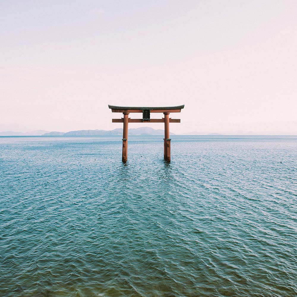 Ein traditionelles rotes japanisches Torii steht allein im ruhigen blauen Meer, mit fernen Bergen am Horizont unter einem hellen, klaren Himmel.