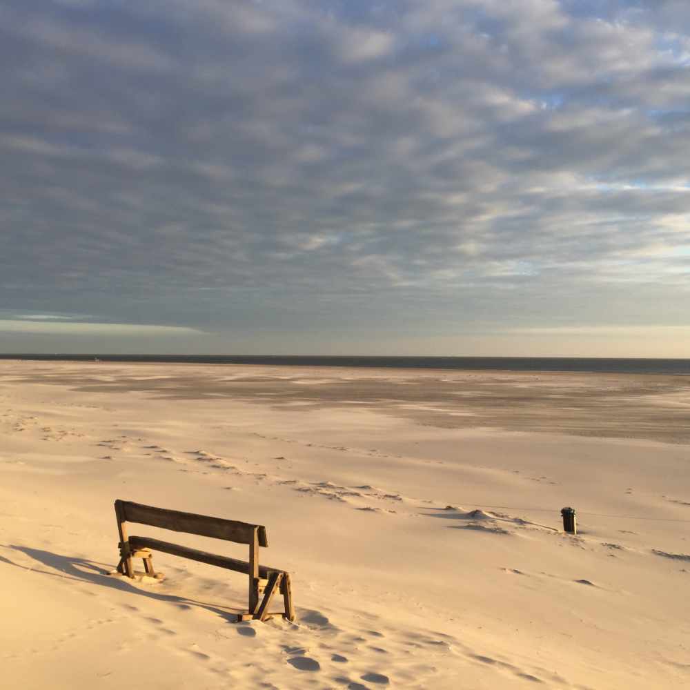 Leere Holzbank im Sand mit Blick auf einen weiten Strand und das Meer unter bewölktem Himmel.